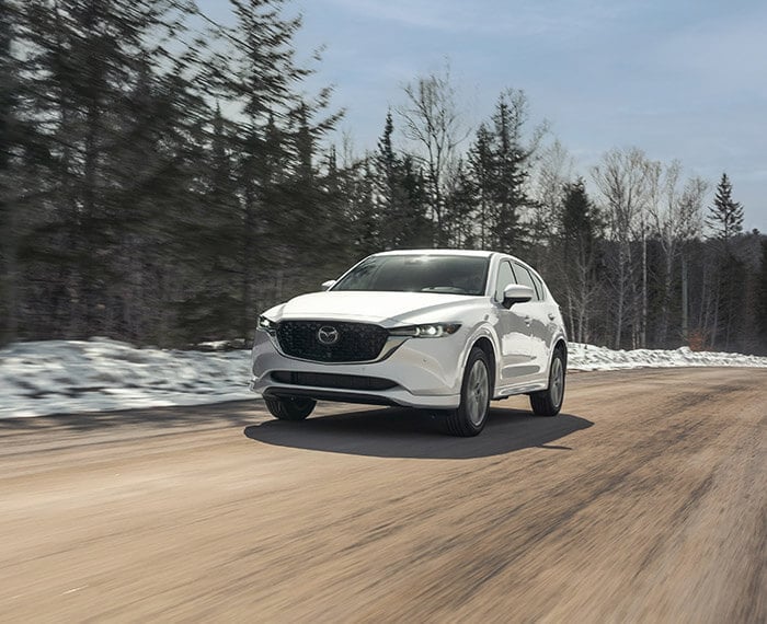 Vue 3/4 avant du Mazda CX-5 2023 Blanc Rhodium métallisé roulant sur un chemin de terre