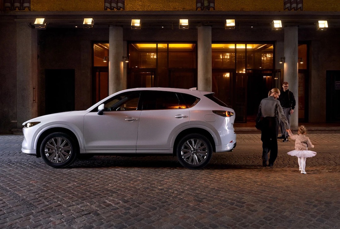 Vue latérale du Mazda CX-5 2023 Blanc Rhodium métallisé garé en ville avec des piétons à l'arrière du VUS.